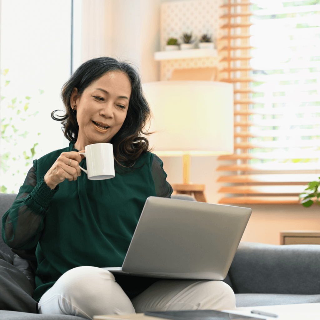 A woman sitting on a couch, a laptop on her lap and a cup of coffee, watching screen with a smile