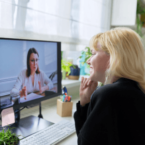 A woman sitting at a desking smiling while talking to a therapist on zoom