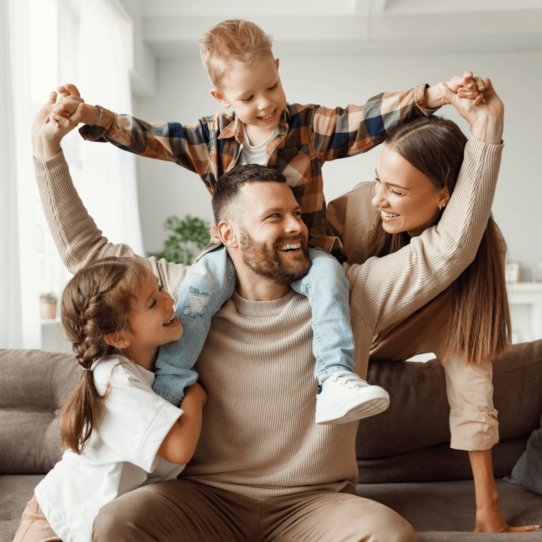 Afather-sitting on a couch, young son on shoulders with daughter and wife either side, all smiling