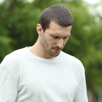 man in 30s in a park, looking down with eyes closed and a slight concerned frown