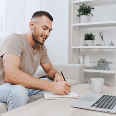 A man taking notes on a coffee table with laptop one and a cup of coffee
